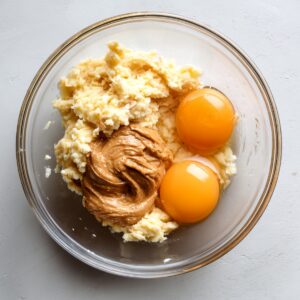 A glass mixing bowl holding mashed banana, two cracked egg yolks, and a dollop of almond butter, ready to be mixed.