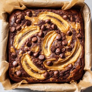 Freshly baked banana–peanut butter swirl brownies studded with chocolate chips, set in a parchment‑lined square pan.