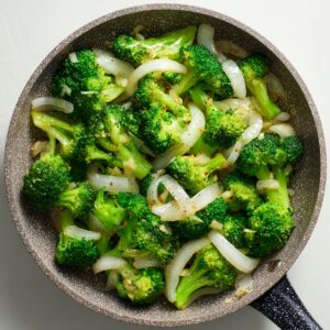 Overhead view of sautéed broccoli florets mixed with thin white onion slices in a speckled frying pan.