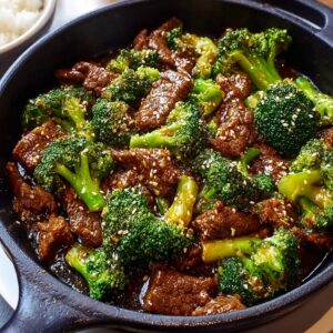 Close-up skillet of Beef and Broccoli Recipe in glossy brown sauce, sprinkled with sesame seeds, rice blurred in background