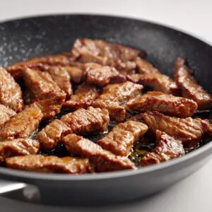 Three‑quarter close-up of seared beef strips glistening with oil in a speckled nonstick pan.