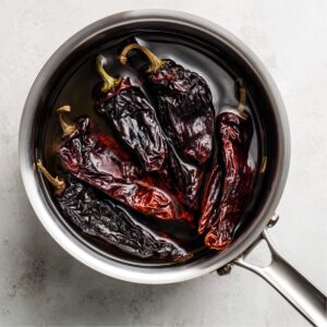Top-down view of dried guajillo and ancho chiles soaking in a stainless steel saucepan filled with warm water, ready to be blended into sauce.