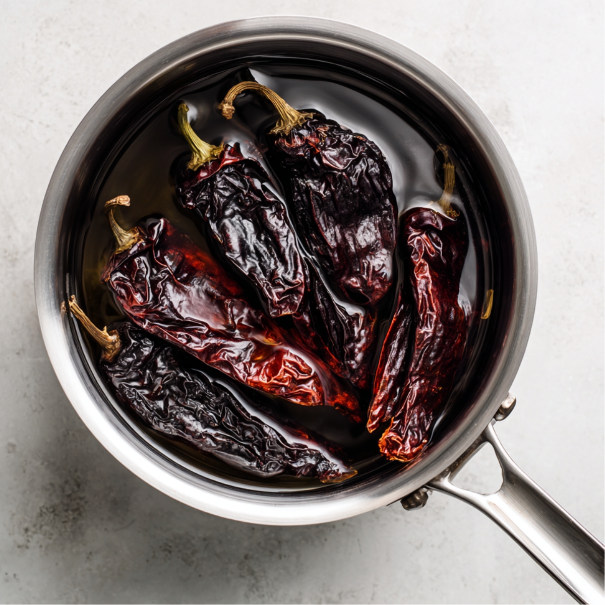 Top-down view of dried guajillo and ancho chiles soaking in a stainless steel saucepan filled with warm water, ready to be blended into sauce.