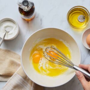 A white ceramic bowl contains two cracked eggs and vegetable oil being whisked together by hand, surrounded by a jar of syrup and a small bowl of salt, for a smooth batter in this chocolate cake recipe.