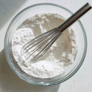 Top‑down view of a clear glass mixing bowl on a white speckled countertop: white all‑purpose flour partially mixed with milk, with a stainless‑steel balloon whisk resting in the bowl; soft shadows fall across the bright workspace