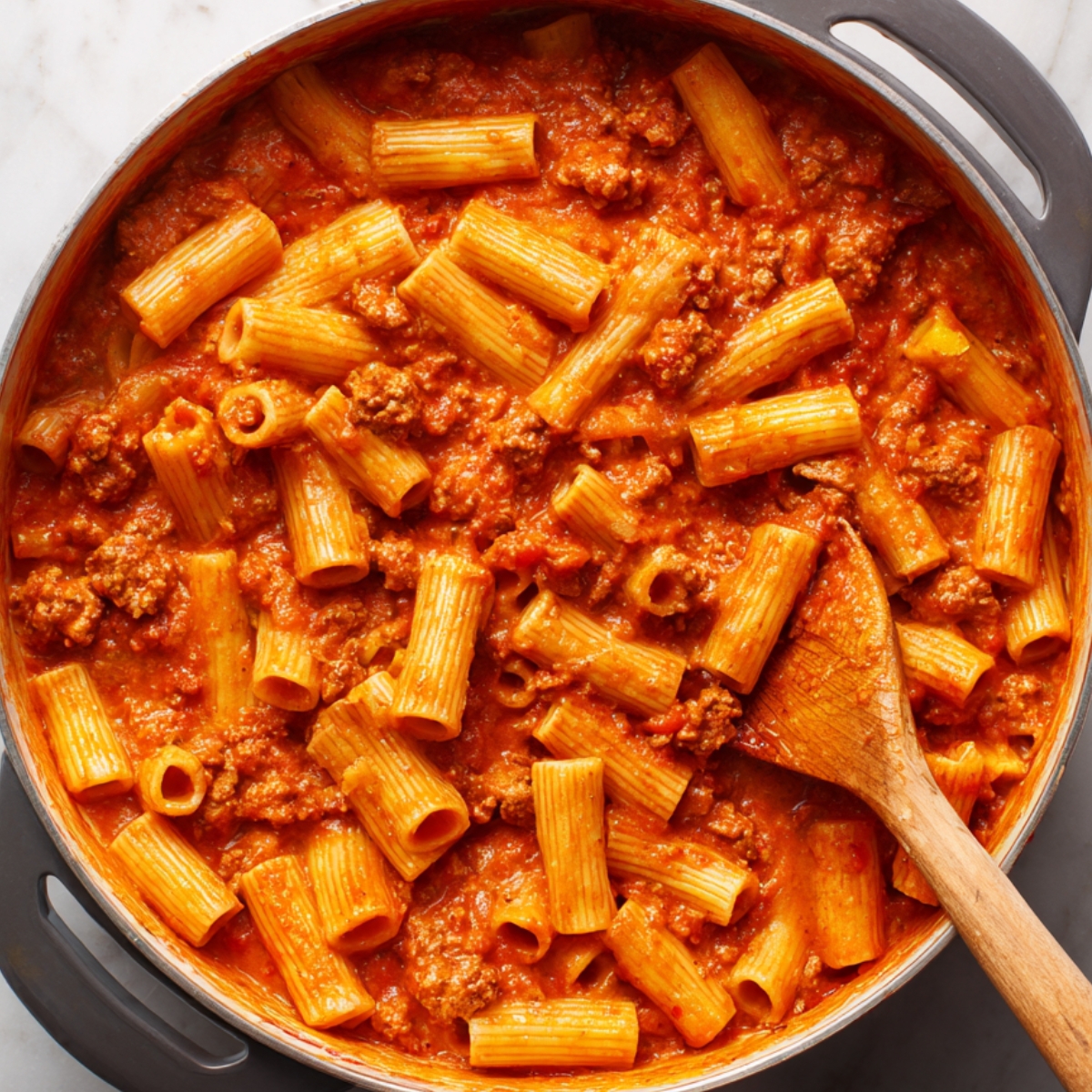 A top‑down view into a wide skillet filled with rigatoni coated in creamy tomato sauce, a wooden spoon stirring the pasta as a metal measuring cup pours in reserved pasta cooking water.