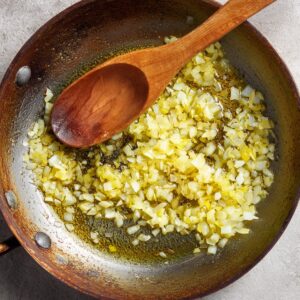Overhead view of finely diced yellow onion sizzling in olive oil in a deep skillet, a wooden spoon lying across the pan.