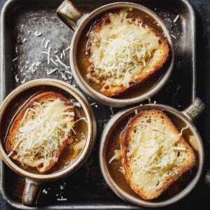 Top-down view of three bowls of French onion soup on a baking sheet, toasted baguette slices covered with shredded cheese.