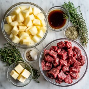 Overhead view of bowls with diced steak, cubed potatoes, butter, Worcestershire sauce, dried herbs, salt, pepper, and fresh thyme/rosemary on marble.