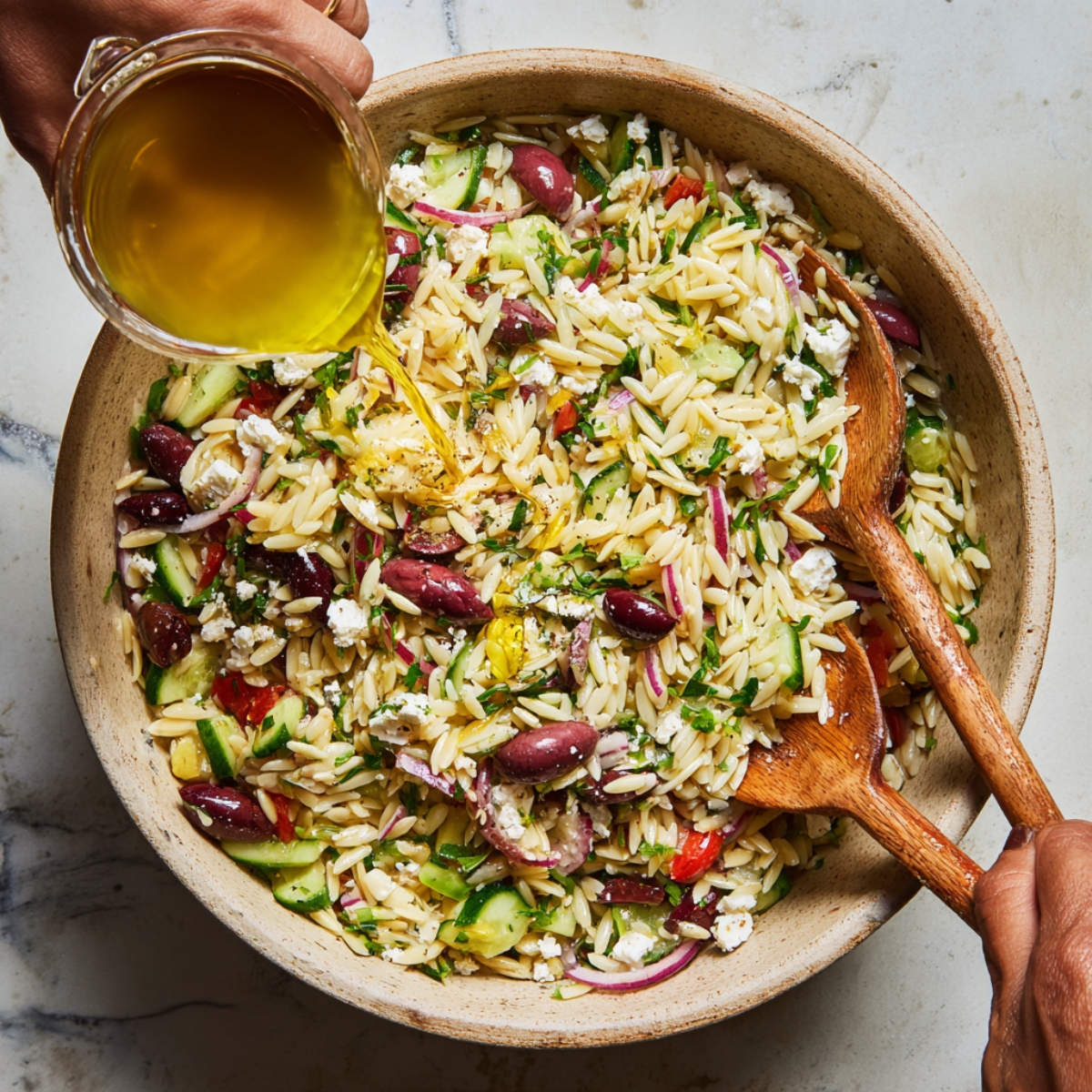 High‑angle photograph of a wooden salad bowl filled with orzo, tomatoes, cucumber, olives, red onion, and feta as golden olive oil is poured over it; two wooden servers rest inside the bowl