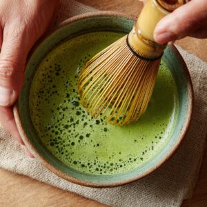 Overhead view of a speckled ceramic bowl as a hand uses a bamboo whisk to vigorously froth vibrant green matcha into a smooth, bubbly mixture.