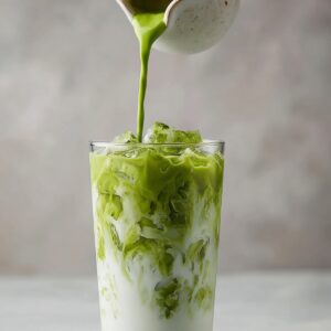 A tall clear glass filled with ice as bright green matcha is poured from a white pitcher, creating swirling patterns through creamy milk.