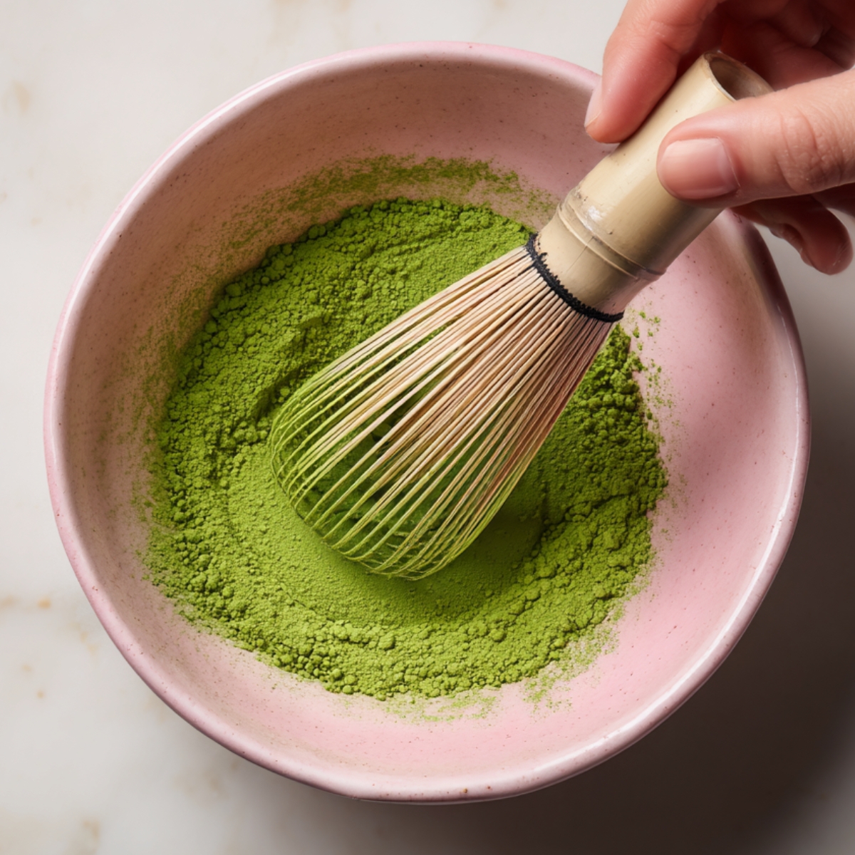 Top‑down shot of a pink bowl holding vivid green matcha powder with a bamboo whisk resting on the rim, ready for whisking.