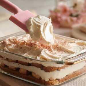 A pink silicone spatula smoothing a thick layer of pale mascarpone cream over a neat first layer of espresso‑soaked ladyfingers in a square glass baking dish, soft kitchen lighting and marble background.