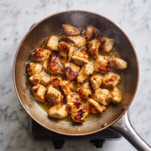 Bird’s‑eye view of bite‑sized chicken pieces searing in oil in a light-colored Dutch oven on an induction burner, developing a golden brown crust.