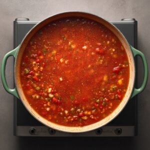 Top‑down shot of an enameled pot on an induction cooktop containing a thick, bubbling tomato‑pepper sauce with visible diced vegetables and Cajun spices