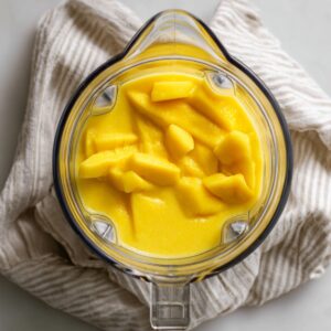 Overhead shot of a clear blender pitcher on a white counter, filled midway with smooth, vibrant yellow mango puree and a few larger mango chunks visible on top, with the blender base and a folded striped kitchen towel beside it.