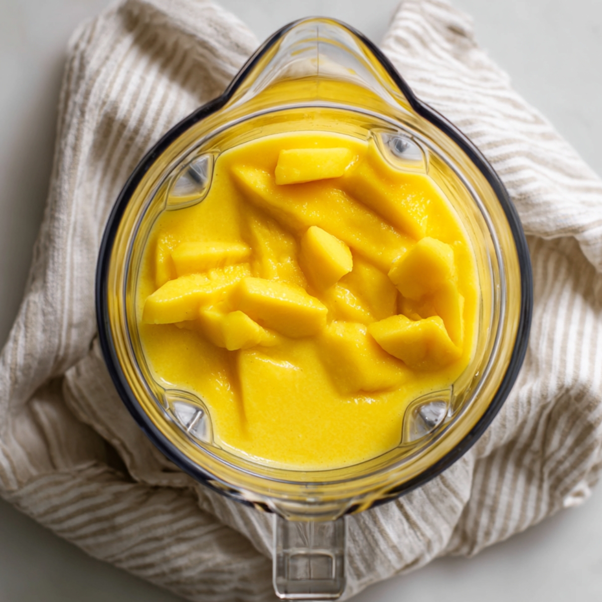 Overhead shot of a clear blender pitcher on a white counter, filled midway with smooth, vibrant yellow mango puree and a few larger mango chunks visible on top, with the blender base and a folded striped kitchen towel beside it.