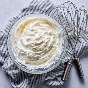 Top‑down shot of a glass bowl on a light, speckled countertop containing freshly whipped cream with soft peaks, next to two metal mixer beaters resting on a striped dish towel.