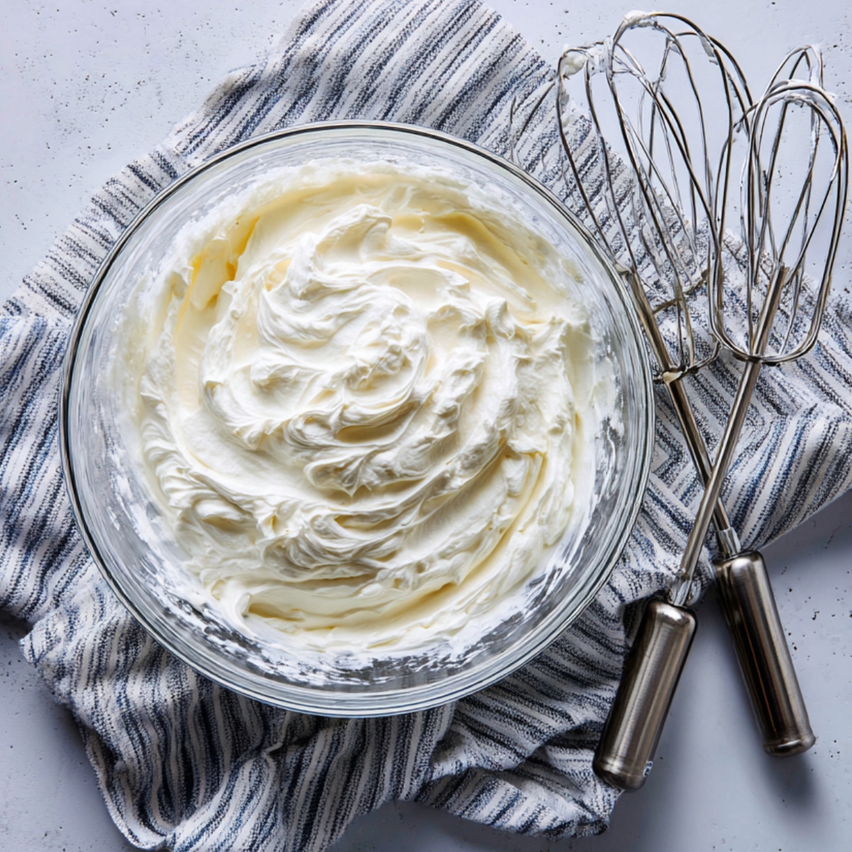 Top‑down shot of a glass bowl on a light, speckled countertop containing freshly whipped cream with soft peaks, next to two metal mixer beaters resting on a striped dish towel.