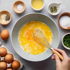 Overhead view of a rustic wooden tabletop scattered with omelette fixings: whole eggs in a wooden bowl, fried eggs in a small dish, sliced mushrooms, wilted spinach, chopped prosciutto, a small bowl of shredded cheese, a cup of olive oil, a bowl of cream, a dish of coarse salt, ground pepper, and dried herbs—all ready for assembly.