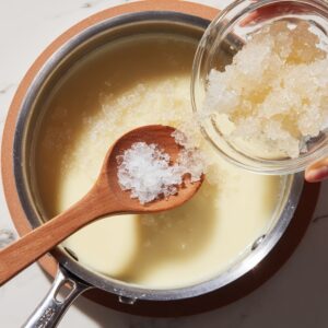 Close‑up overhead of a saucepan on a trivet with steaming cream, a wooden spoon holding a mound of fully bloomed, translucent gelatin crystals about to be added from a clear glass bowl.
