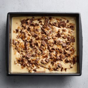 Overhead view of a white rectangular pan filled with glossy brownie batter swirled with caramel and studded with chopped candy pieces, ready to bake.