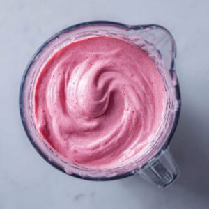 Top‑down shot of a smooth, velvety pink strawberry mixture swirling inside a transparent blender pitcher, with tiny flecks of fruit visible against a soft gray background