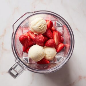 Overhead view into a clear blender pitcher on a light marble surface, filled with bright red hulled strawberries and three generous scoops of pale‑yellow vanilla ice cream