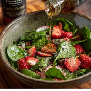 Overhead shot of a glass bowl filled with a speckled, whisked dressing: a glossy mixture of apple cider vinegar, honey, Dijon mustard, olive oil, lemon juice, and visible poppy seeds, with a metal whisk resting inside.