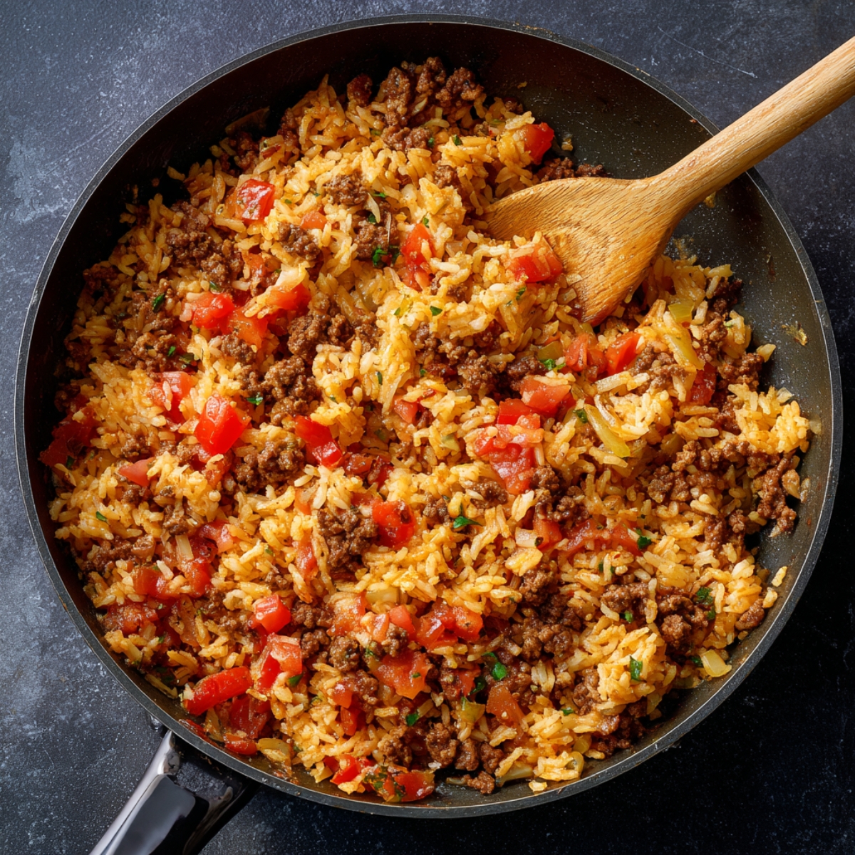 Close‑up overhead of a black skillet on a dark countertop, showing browned ground beef mixed with white rice, diced tomatoes, and onions in a light sauce, with a wooden spoon resting inside.