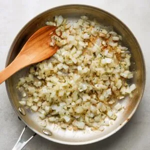 A flat‑lay of a nonstick skillet on a pale marble surface, filled with translucent sautéed diced onions that are just beginning to brown, with a wooden spatula at the edge stirring them.