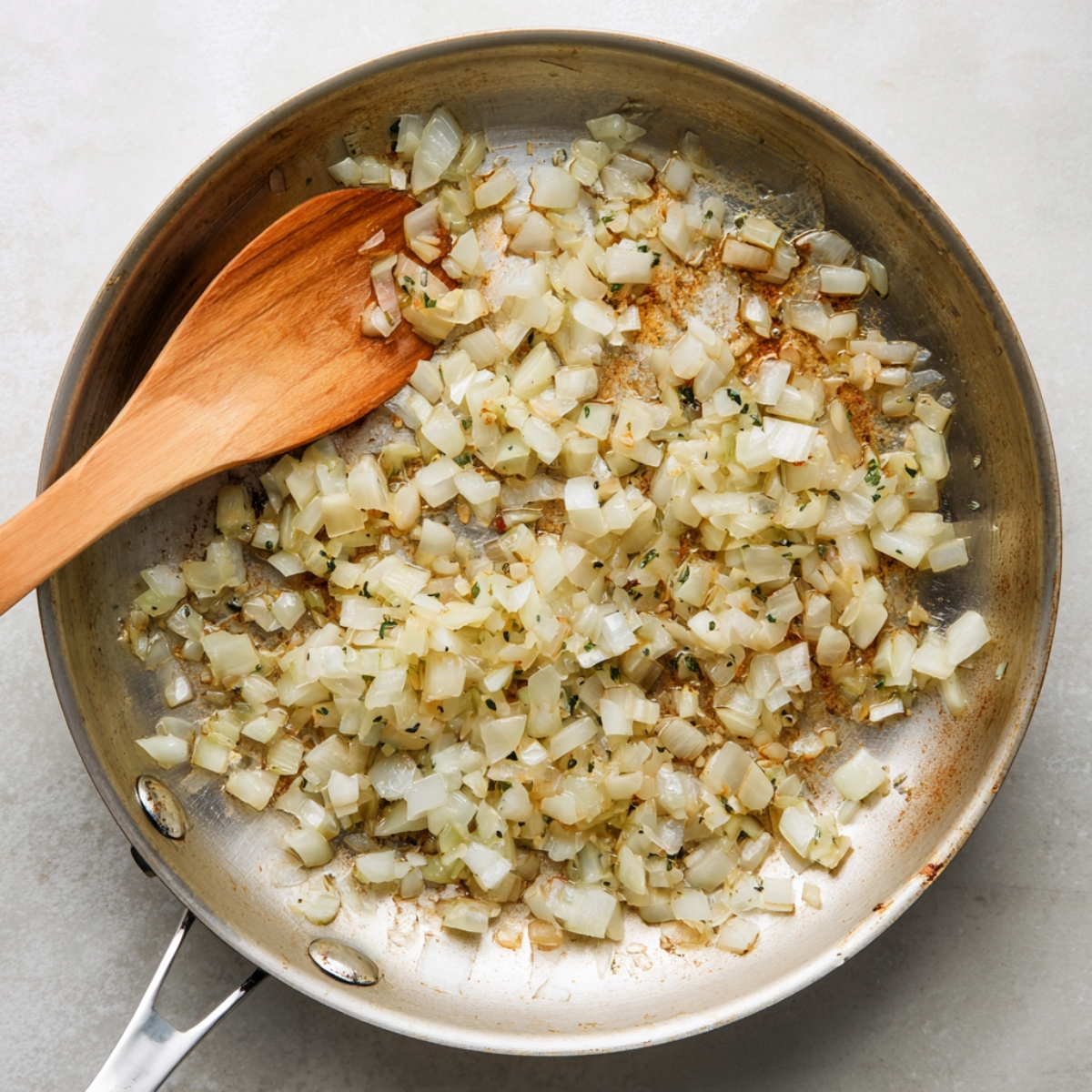 A flat‑lay of a nonstick skillet on a pale marble surface, filled with translucent sautéed diced onions that are just beginning to brown, with a wooden spatula at the edge stirring them.