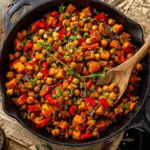 A top‑down view of a black cast‑iron skillet on a rustic wooden table lined with vintage newspaper, filled with vibrant sweet potato and chickpea hash studded with red pepper pieces and fresh thyme sprigs, a wooden spoon resting in the pan.