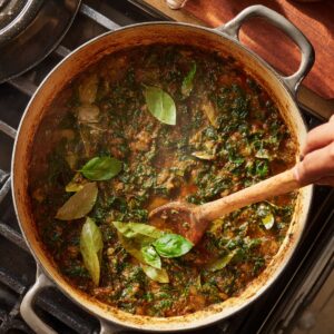 A large Dutch oven filled with a rich stew being stirred with a wooden spoon, topped with fresh bay leaves and basil, simmering on a stovetop.