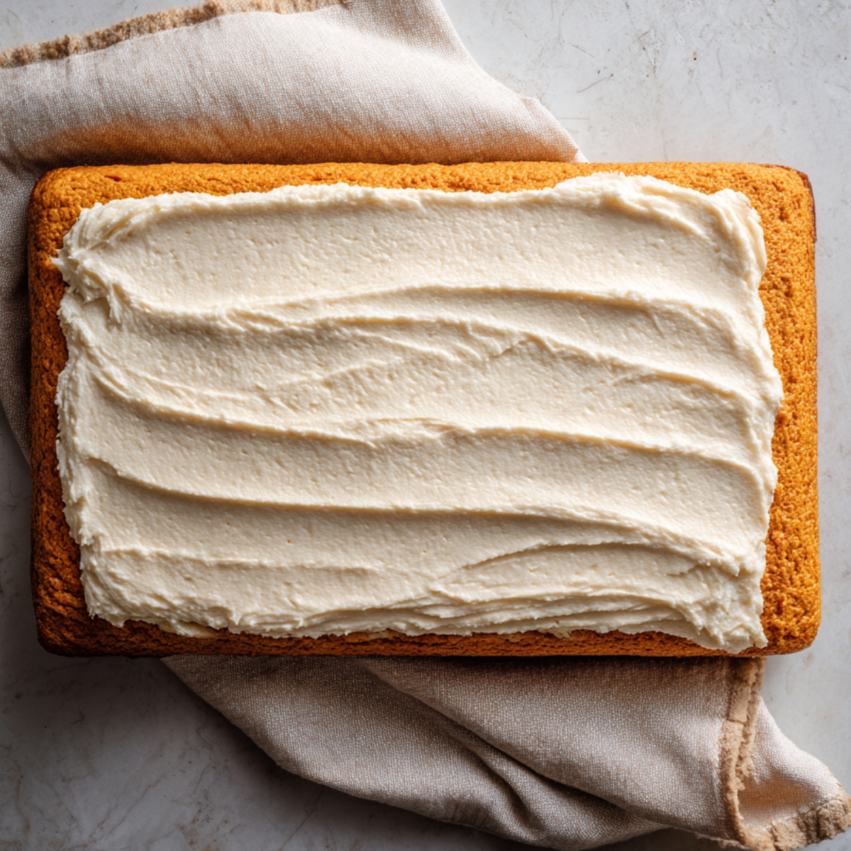 Overhead pumpkin sheet cake on a towel, spread with an even layer of cream cheese filling, ready to roll.