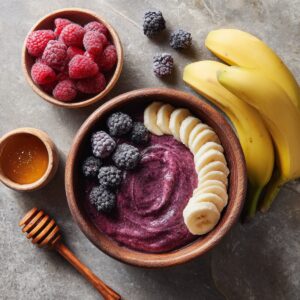 A wooden bowl filled with purple acai mixture topped with banana slices and blackberries, with a small bowl of honey and raspberries placed beside it.
