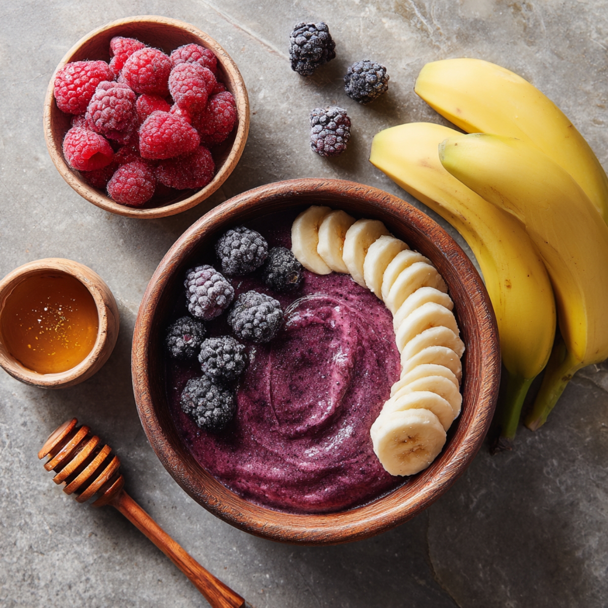 A wooden bowl filled with purple acai mixture topped with banana slices and blackberries, with a small bowl of honey and raspberries placed beside it.