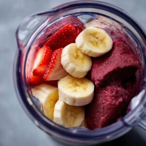 A blender jar containing slices of strawberries, bananas, and acai puree, ready to be blended into an acai bowl.