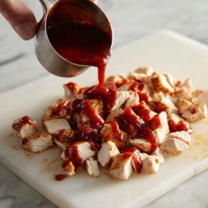 A close-up of diced grilled chicken breast on a white plastic cutting board, being drizzled with thick, glossy BBQ sauce from a metal measuring cup. The chicken pieces are cooked and lightly browned, and the BBQ sauce is rich and dark red. The setting is a white marble countertop with natural light, showing a casual kitchen prep scene.
