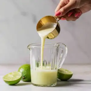 A hand with coral-colored nail polish pours sweetened condensed milk from a shiny gold measuring cup into a clear pitcher with pale lime juice. Limes are arranged casually around the base. All scenes should appear 100% real, naturally lit, and homemade."