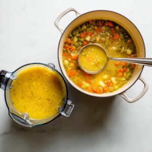 A pot of just-cooked vegetable soup filled with colorful diced carrots, potatoes, and celery, alongside a blender half-filled with the smooth, golden blended version. A metal ladle rests in the pot, showing the transition from chunky to creamy soup.