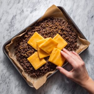 Overhead view of a small metal sheet pan lined with crumpled brown parchment paper, packed with freshly cooked, crumbled ground beef. A neat stack of six bright orange cheddar cheese squares sits atop the meat, while a right hand reaches in to place another slice at the corner. The pan rests on a white marble countertop with subtle gray veining, and the scene is lit by soft, natural light that highlights the meat’s glistening juices and the cheese’s glossy surface.