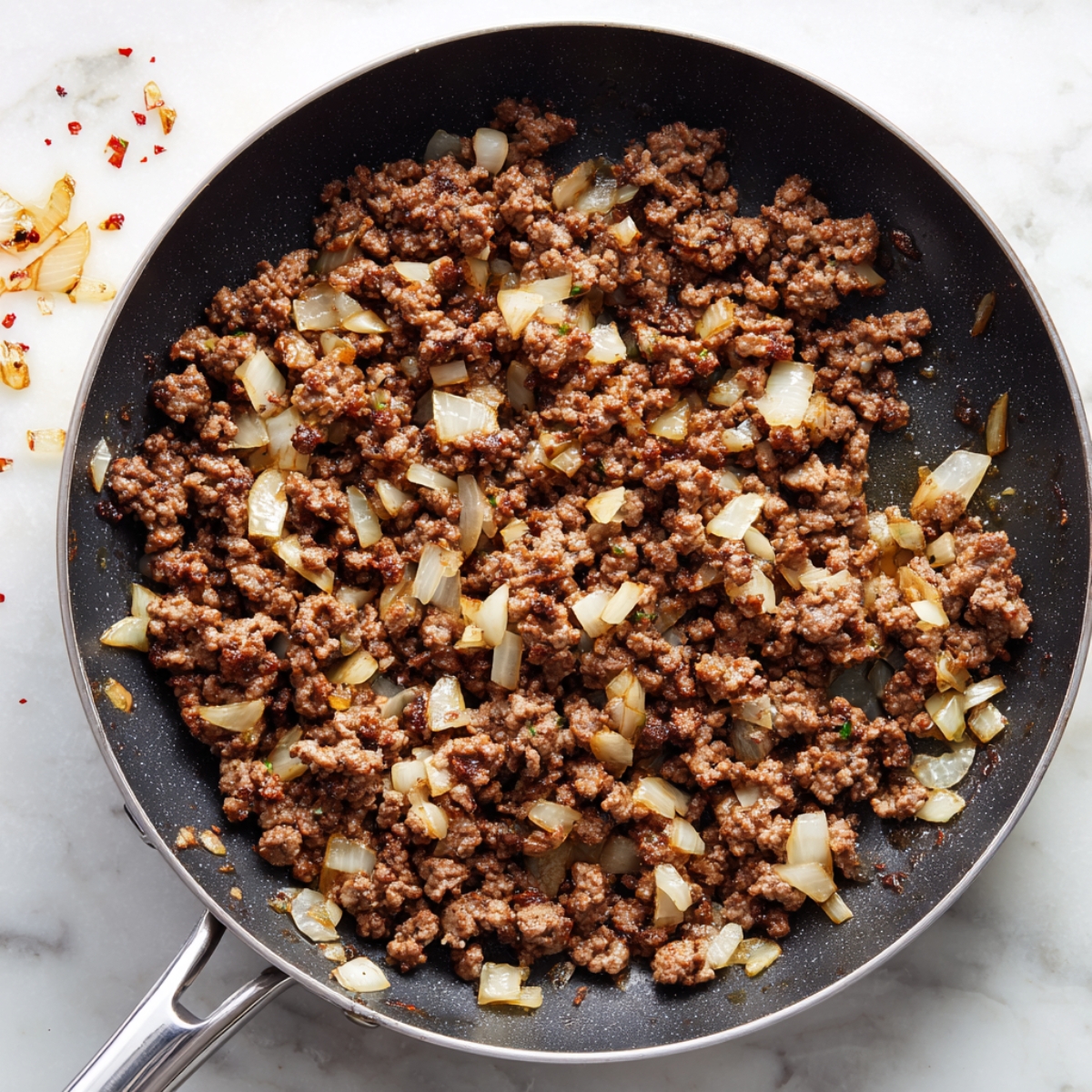 Overhead view of a black nonstick skillet with a stainless-steel handle, filled with freshly browned, crumbled ground beef sautéed together with translucent, golden-edged diced onions. A few scattered red pepper flakes cling to the beef and tiny pools of glossy cooking oil catch the light. The skillet rests on a white marble countertop dusted here and there with stray onion bits and chili flakes, evoking a homey, just-cooked feel.