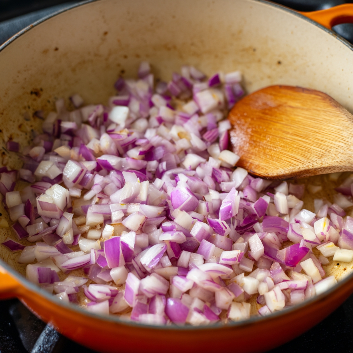 Orange enameled pot on the stove containing diced red onion being stirred with a wooden spoon as it softens