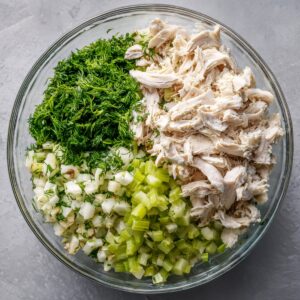Glass mixing bowl filled with shredded cooked chicken, diced celery, finely chopped fresh dill, and chopped green onions ready to be combined.”