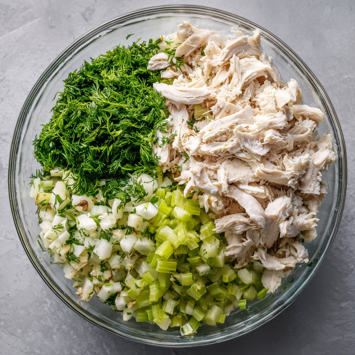 Glass mixing bowl filled with shredded cooked chicken, diced celery, finely chopped fresh dill, and chopped green onions ready to be combined.”