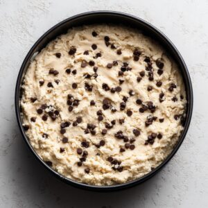 Overhead shot of a black springform pan filled with unbaked chocolate chip cookie dough pressed evenly into the bottom. The pale marble countertop beneath and gentle natural light emphasize the dough’s smooth texture and scattered semi-sweet chips