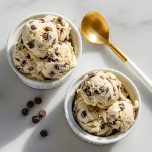 Two white ceramic ramekins sit on a pale marble surface, each piled high with three scoops of homemade edible cookie dough flecked with semi-sweet chocolate chips. A gold-tipped spoon rests beside the ramekins, and a few loose chips are scattered around, all bathed in soft, natural light that highlights the dough’s creamy texture and the spoon’s warm metallic sheen.