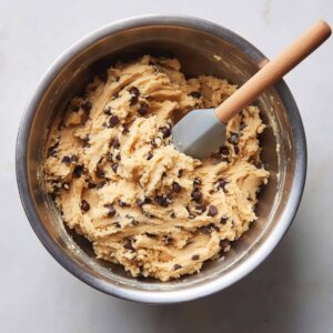 A stainless-steel mixing bowl on a marble counter holds thick, freshly mixed edible cookie dough teeming with chocolate chips. A wooden-handled, gray silicone spatula is buried in the dough, its blade coated and pulling up a textured scoop. The even, diffused lighting brings out the dough’s rough peaks and the spatula’s smooth surface.